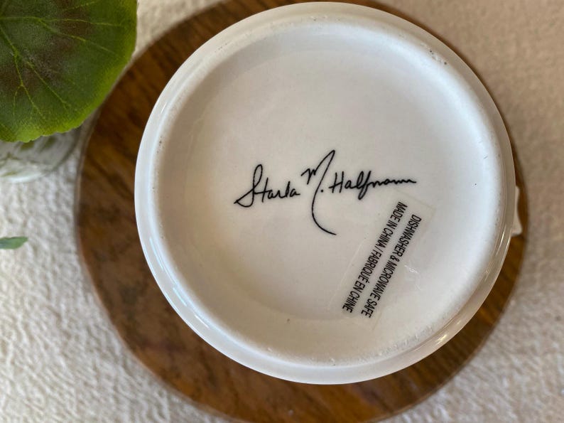 Puede incluir: Base de una taza de cer&aacute;mica blanca con la firma "Starla M. Halfmann" en escritura negra. Una etiqueta dice "Apto para lavavajillas y microondas Made in China / Fabriqu&eacute; en Chine."