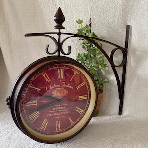 May include: A double-sided, antique-style wall clock with a burgundy face and Roman numerals. The clock features the text "Chateau Joullian Grand Vin Bordeaux" and is mounted on a decorative, dark metal bracket. A small potted plant is visible behind the clock.