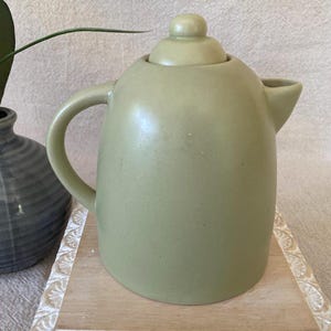 May include: A sage green ceramic teapot with a rounded body, a curved handle, and a small spout. The lid has a rounded knob. The teapot sits on a light-colored wooden tray. A dark gray vase and a plant are in the background.