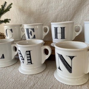 May include: White ceramic mugs with black letters, including C, E, H, K, L, M, and N. Each mug has a handle and a pedestal base. The letters are in a decorative font. A small evergreen tree is in the background.