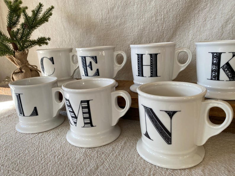 May include: White ceramic mugs with black letters, including C, E, H, K, L, M, and N. Each mug has a handle and a pedestal base. The letters are in a decorative font. A small evergreen tree is in the background.