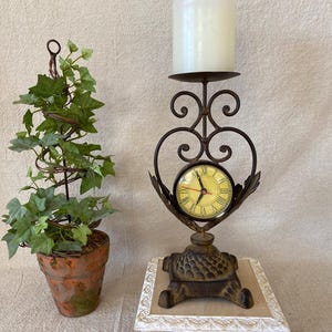 May include: An ornamental candle holder with a clock and a potted plant. The candle holder is made of dark metal with a heart-shaped design and a round clock. The potted plant has green leaves and a spiral metal frame.