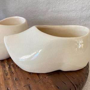 May include: Two off-white ceramic clogs, a traditional Dutch design, sit on a wooden surface. The clogs have a glossy finish and a hollow interior, suggesting they are decorative planters or small containers.