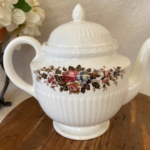 May include: White ceramic teapot with a lid and a floral design. The teapot has a ribbed body and a curved handle. The floral design includes pink, red, blue, and brown flowers. The teapot is on a wooden surface.