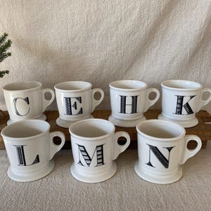 May include: Seven white ceramic mugs with black letters C, E, H, K, L, M, and N. Each mug has a handle and a pedestal base. The mugs are arranged on a wooden surface.