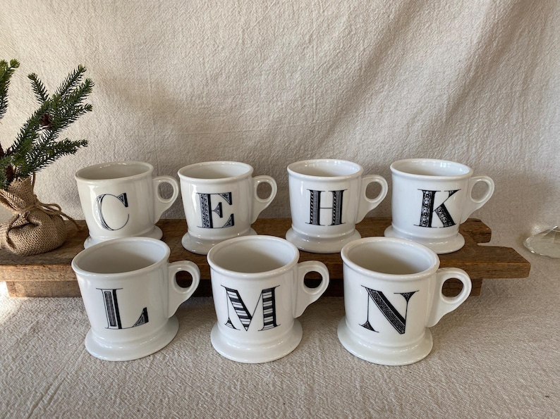 May include: Seven white ceramic mugs with black letters C, E, H, K, L, M, and N. Each mug has a handle and a pedestal base. The mugs are arranged on a wooden surface.