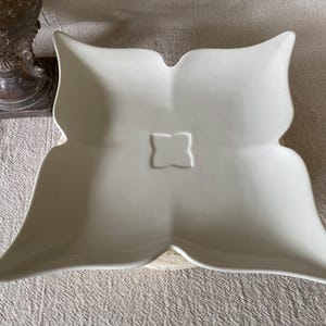 May include: A white, four-lobed ceramic serving dish with a square, raised design in the center. The dish has a smooth, glossy finish and is set on a neutral-colored surface. The dish's shape resembles a stylized flower.