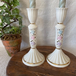 May include: Pair of off-white ceramic candlesticks with gold trim and floral cutouts. Each candlestick holds a gray candle and features a fluted top and base. A small potted plant with green leaves sits to the left.
