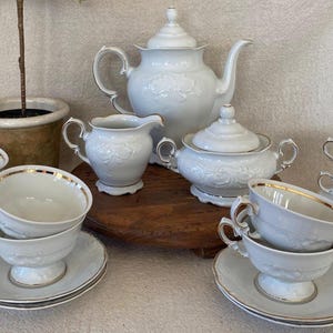 May include: A white porcelain tea set with gold trim, including a teapot, sugar bowl, creamer, and several teacups and saucers. The set has a floral embossed design. The tea set is arranged on a wooden surface.