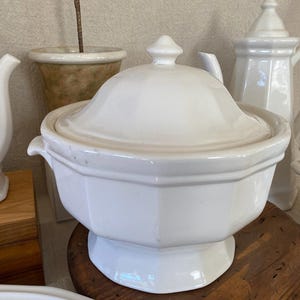 May include: A white ceramic soup tureen with a lid, featuring a pedestal base and side handles. The tureen has a geometric design and is part of a collection of white ceramic serving pieces. A white teapot is also visible.