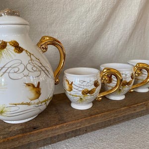May include: White ceramic tea set with gold accents, featuring a teapot and four teacups. The set is decorated with illustrations of birds, pinecones, and delicate gold lines. The teapot has a decorative gold lid.