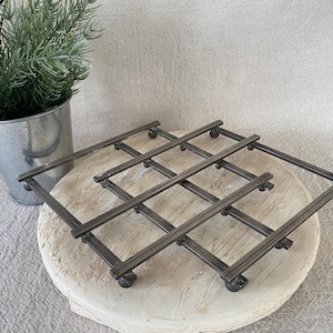 May include: A dark gray metal trivet with a grid design, resting on a light-colored, round wooden surface. The trivet has small, rounded feet. A small metal bucket with greenery is in the background.