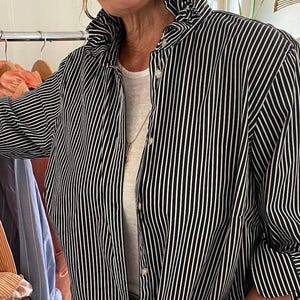 Peut inclure: Une chemise boutonn&eacute;e &agrave; rayures verticales noires et blanches dot&eacute;e d'un col haut &agrave; volants, de manches longues et d'une patte de boutonnage sur le devant. La chemise est port&eacute;e sur un sous-v&ecirc;tement blanc uni, en tissu structur&eacute; et net.