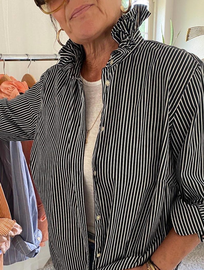 Peut inclure: Une chemise boutonn&eacute;e &agrave; rayures verticales noires et blanches dot&eacute;e d'un col haut &agrave; volants, de manches longues et d'une patte de boutonnage sur le devant. La chemise est port&eacute;e sur un sous-v&ecirc;tement blanc uni, en tissu structur&eacute; et net.