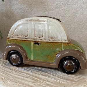 May include: A ceramic car-shaped bank in shades of green, brown, and white. The vintage-style vehicle has a rounded roof and wheels, with a slot on top for inserting coins. The bank is placed on a light-colored, textured surface.