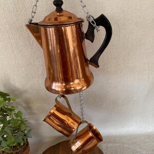 May include: A vintage copper coffee pot with a black handle and lid, suspended by a chain. Two matching copper mugs hang below. The pot and mugs have a shiny, reflective surface, suggesting they are well-maintained.
