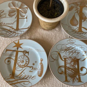 May include: Five decorative plates with gold accents and lettering. Each plate features a different design: a flower, a pear, a pineapple, a clock, and a letter. The plates have a light blue background and gold rims.