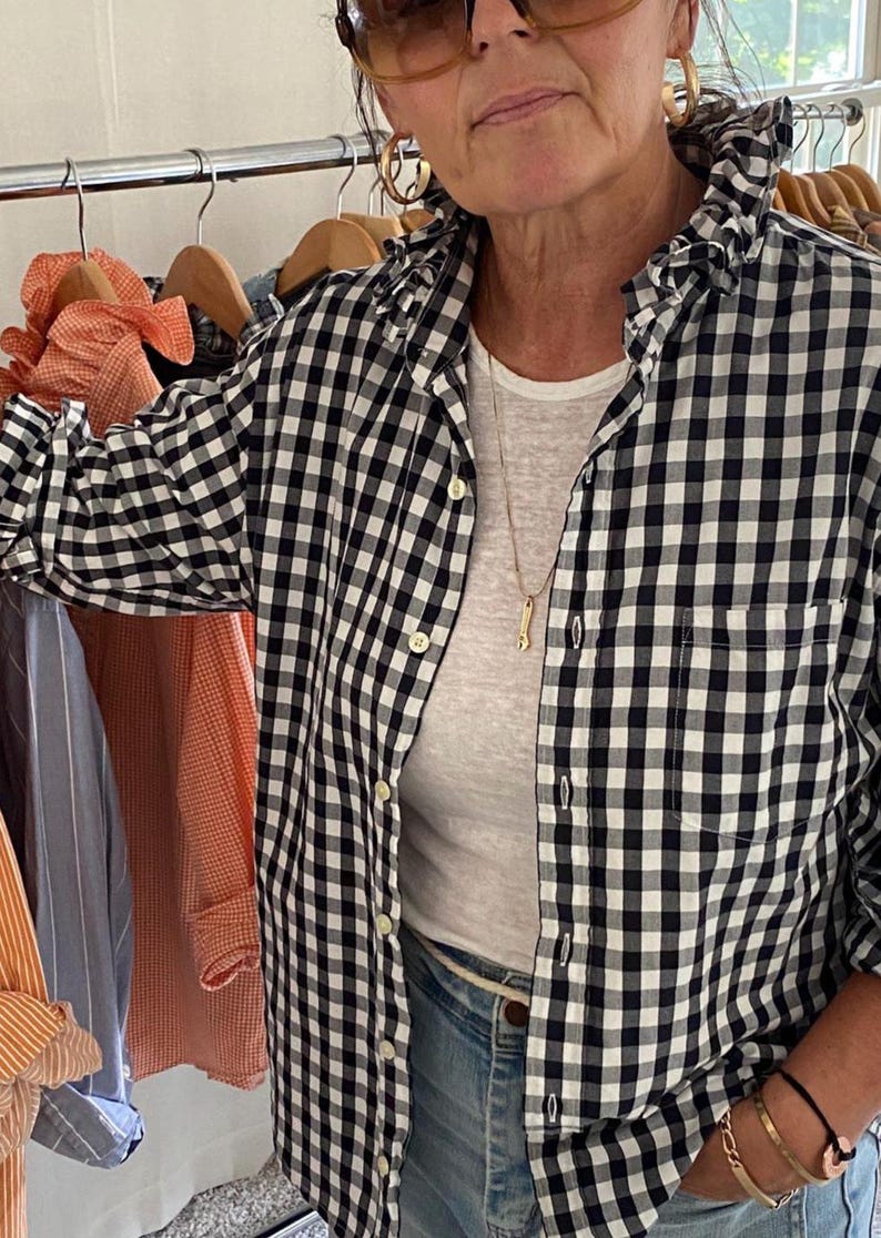 Peut inclure: Une chemise boutonn&eacute;e &agrave; carreaux vichy noirs et blancs avec un col &agrave; volants, des manches longues et une poche poitrine. La chemise est port&eacute;e sur un t-shirt blanc uni avec un jean bleu, un collier dor&eacute; et des boucles d'oreilles cr&eacute;oles dor&eacute;es.