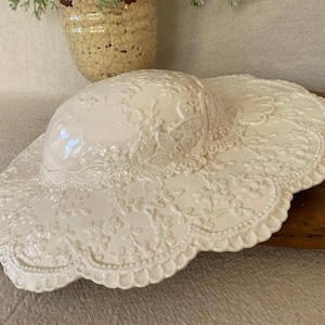 May include: A white ceramic hat with a wide brim, featuring an embossed floral and lace design. The hat has a glossy finish and a scalloped edge. Decorative item.