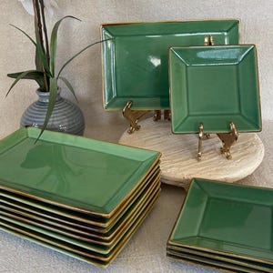 May include: A collection of green ceramic serving dishes with gold trim. The set includes rectangular and square plates, some stacked, and others displayed on decorative stands. A vase with a plant is in the background.