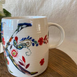 Puede incluir: Taza de cer&aacute;mica blanca con un gran dise&ntilde;o floral en rojo, azul y verde. La taza tiene un asa curva y est&aacute; sobre una superficie de madera. El dise&ntilde;o floral incluye la letra "K".