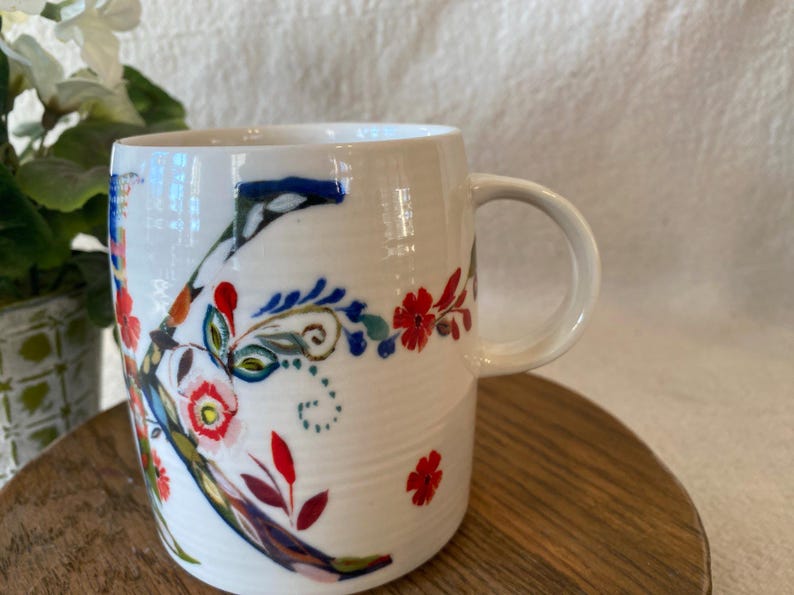 Puede incluir: Taza de cer&aacute;mica blanca con un gran dise&ntilde;o floral en rojo, azul y verde. La taza tiene un asa curva y est&aacute; sobre una superficie de madera. El dise&ntilde;o floral incluye la letra "K".