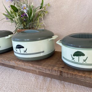 May include: Three vintage food warmers with gray lids and handles. The off-white containers have a black graphic of an umbrella and chairs. The warmers sit on a wooden surface with a plant in the background.