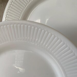 May include: Close-up of two white ceramic plates. The plates have a glossy finish and a ribbed, textured border. The plates are stacked, with one partially visible behind the other. The plates are clean and simple, suitable for everyday use.