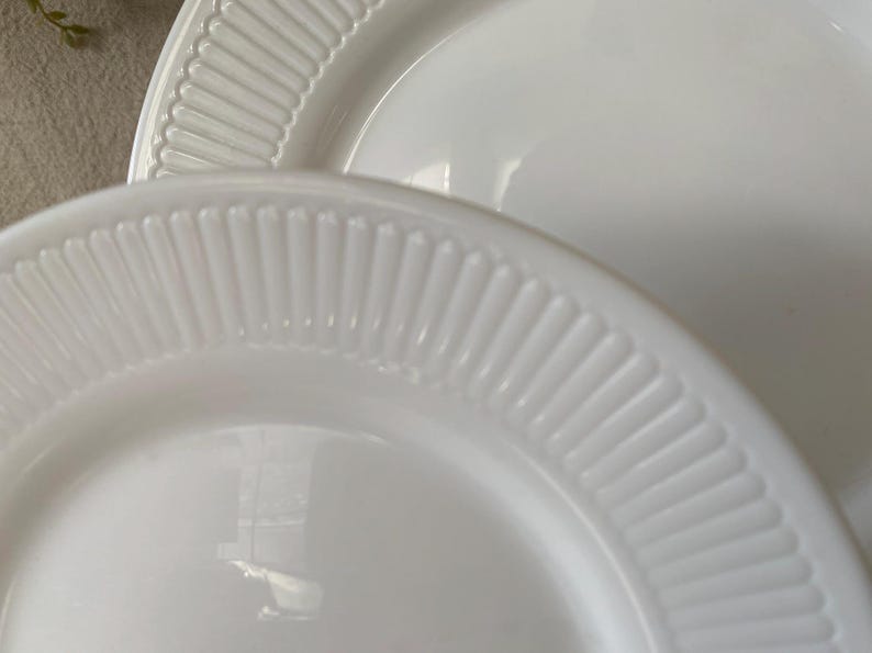 May include: Close-up of two white ceramic plates. The plates have a glossy finish and a ribbed, textured border. The plates are stacked, with one partially visible behind the other. The plates are clean and simple, suitable for everyday use.