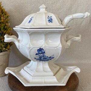 May include: A white ceramic soup tureen with a lid and a matching ladle. The tureen features blue floral designs and is set on a matching base. The handles are ornate and curved.