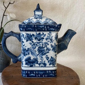May include: A blue and white ceramic teapot with a square body and a curved handle. The teapot features a floral pattern and a lid with a finial. The spout is angular, and the overall design is traditional.