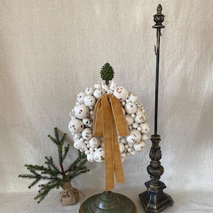May include: A decorative Christmas wreath made of white spheres with red accents and a gold ribbon. A small evergreen tree in a burlap sack and a tall, ornate black candle holder are also present.