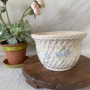 May include: A cream-colored ceramic planter featuring a raised basket-weave texture and embossed blue floral accents. The round vessel has a flared rim and a tapered base, showcasing a detailed lattice pattern across the exterior surface.