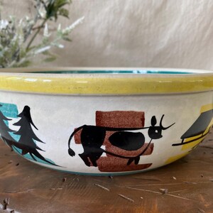 May include: A ceramic bowl with a hand-painted design featuring a black cow, trees, and a yellow rim. The bowl has a teal handle and a white base with a teal interior. The bowl is on a wooden surface.