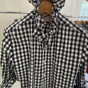 Peut inclure: Une chemise &agrave; manches longues boutonn&eacute;e avec un motif &agrave; carreaux vichy noir et blanc. La chemise pr&eacute;sente un col &agrave; volants pro&eacute;minent et une patte de boutonnage sur le devant. Le tissu arbore un motif &agrave; carreaux classique sur tout le v&ecirc;tement.