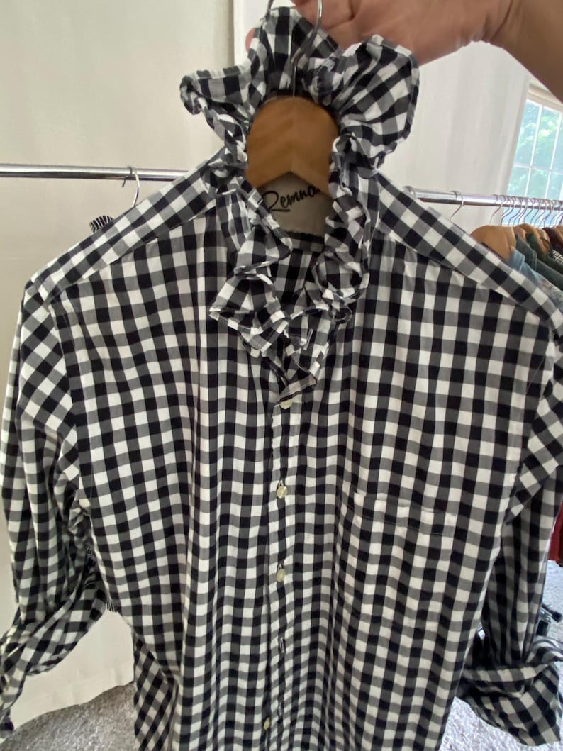 Peut inclure: Une chemise &agrave; manches longues boutonn&eacute;e avec un motif &agrave; carreaux vichy noir et blanc. La chemise pr&eacute;sente un col &agrave; volants pro&eacute;minent et une patte de boutonnage sur le devant. Le tissu arbore un motif &agrave; carreaux classique sur tout le v&ecirc;tement.