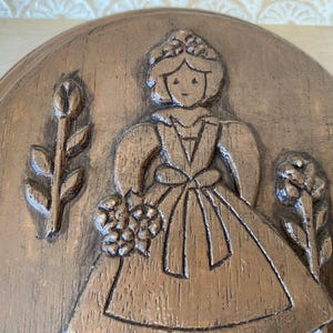 May include: A round, bronze-colored relief carving featuring a woman in a dress holding flowers, flanked by floral and plant motifs. The carving has a textured, wood-like appearance, with detailed lines and shadows.