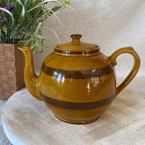 May include: A round ceramic teapot featuring a glossy mustard yellow glaze with two horizontal dark brown stripes encircling the body. The teapot includes a matching lid with a small knob handle, a curved spout, and a sturdy side handle.