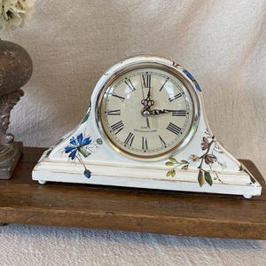 May include: A white mantel clock with a distressed finish, featuring a round face with black Roman numerals and ornate black hands. The clock body is decorated with painted blue and brown floral motifs and includes the text Quartz Westminster Chime.
