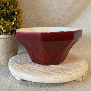 May include: A deep red, hexagonal bowl with a smooth, wide rim and a textured, repeating leaf-like pattern on the exterior sides. The interior of the bowl is a contrasting off-white color, and the overall shape is tapered toward the base.
