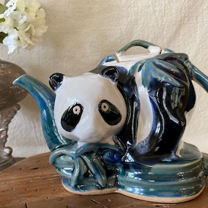 May include: A ceramic teapot featuring a panda bear design. The teapot is primarily white, black, and shades of blue. The panda's face is on the front, with the spout and handle in a bamboo-like design. The teapot is sitting on a wooden surface.
