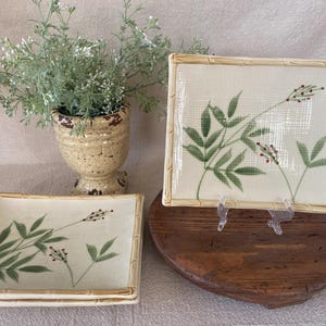 May include: Two square ceramic plates with a bamboo-style border and a painted design of green leaves and red berries. A small, decorative vase with a plant is in the background. The plates are on a wooden surface.