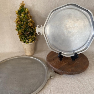 May include: Two vintage metal serving trays. One is oval, the other is octagonal with handles. A small potted plant with green and yellow leaves is in the background. The trays are silver-colored and appear to be made of metal.