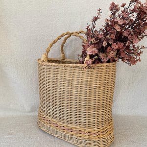 May include: A woven wicker basket with a handle, filled with pink flowers. The basket has a rectangular shape with a rounded bottom and a decorative pink stripe near the base. The handles are made of twisted wicker.