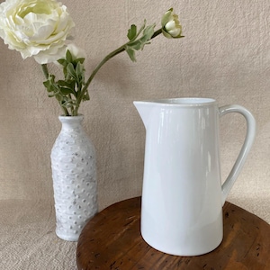 May include: A white ceramic pitcher with a handle and spout sits on a wooden surface. A white textured vase with a flower arrangement is to the left. The flowers are white and green.