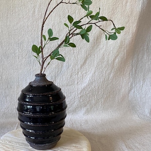 May include: A black ceramic vase with a textured, ribbed design. The vase holds a branch with green leaves. The vase is on a light-colored, round surface against a neutral background.