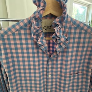Peut inclure: Une chemise &agrave; manches longues boutonn&eacute;e avec un motif &agrave; carreaux vichy rose et bleu. La chemise pr&eacute;sente un col &agrave; volants pro&eacute;minent et une poche poitrine unique. Une &eacute;tiquette int&eacute;rieure blanche avec le texte partiel Rem est visible au col.