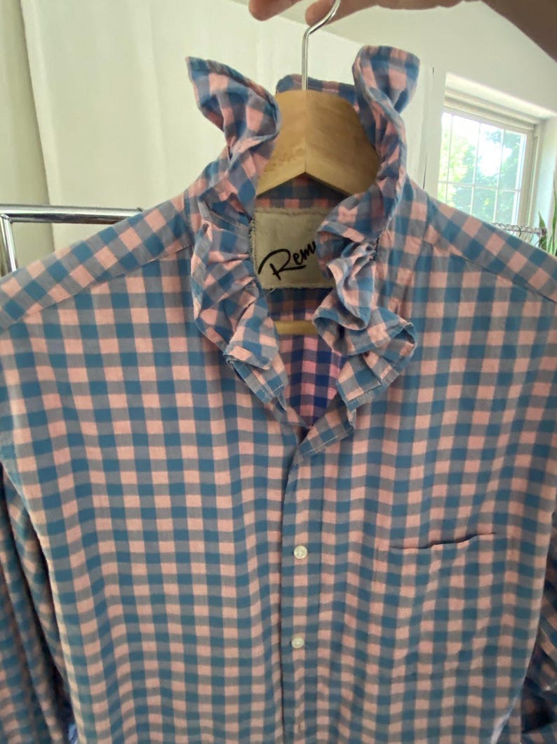Peut inclure: Une chemise &agrave; manches longues boutonn&eacute;e avec un motif &agrave; carreaux vichy rose et bleu. La chemise pr&eacute;sente un col &agrave; volants pro&eacute;minent et une poche poitrine unique. Une &eacute;tiquette int&eacute;rieure blanche avec le texte partiel Rem est visible au col.