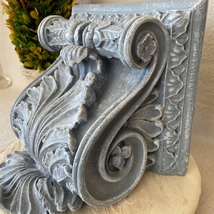 May include: A detailed, gray-blue decorative architectural corbel with ornate scroll and leaf designs. The corbel has a square back and a curved, sculpted front, showcasing classical elements. The piece is likely made of a durable material, suitable for interior design.