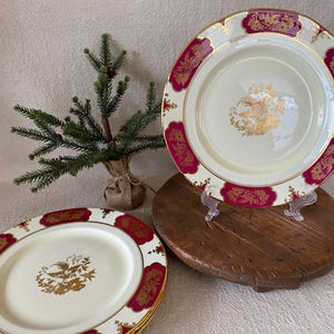 May include: Cream-colored plates with a gold and burgundy floral design. One plate is displayed on a clear stand, while others are stacked. A small evergreen tree in a burlap sack is in the background. The plates have a gold rim.